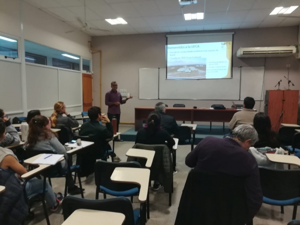 Imagem de um pesquisador da UFCA apresentando seu trabalho em uma sala onde ele está em pé apontando para um slide e algumas pessoas o assistindo sentadas em cadeiras de sala de aula