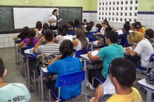 Imagem de uma sala de aula com alunos sentados em suas cadeiras e uma professora a frente ao lado da lousa com um livro na mão