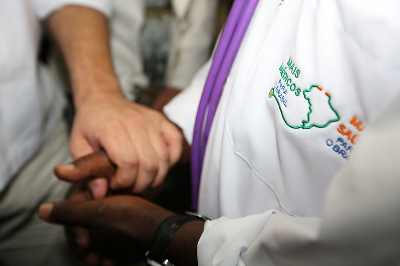Médico de jaleco, com logotipo do Mais Médicos, segurando a mão de um paciente