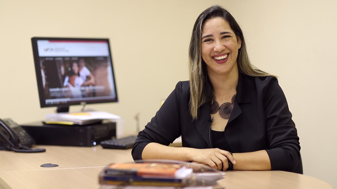 Mulher sorrindo para a câmera, com os braços apoiados na mesa, com um computador atrás dela