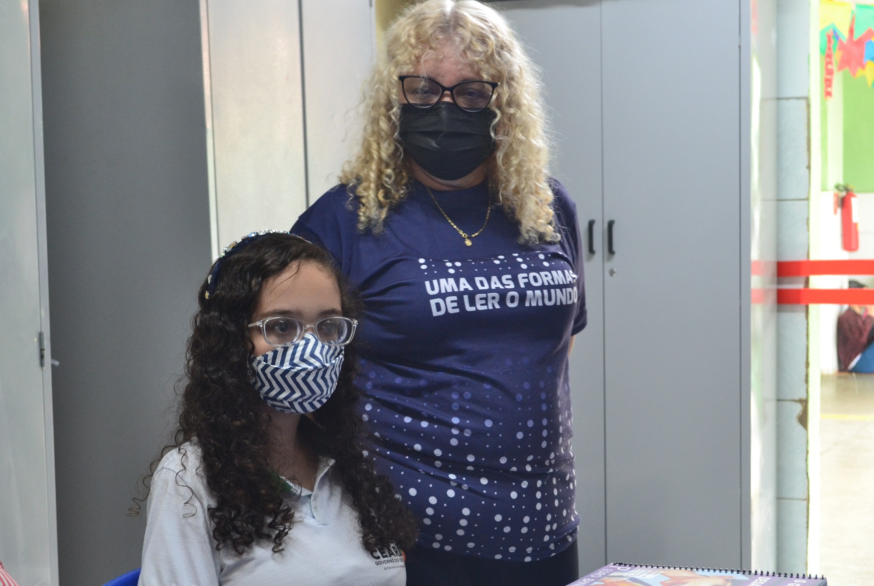 Imagem de duas pessoas. Uma jovem está sentada, ela tem cabelo cacheado loiro, utiliza óculos e veste uma blusa branca ao seu lado em pé há uma mulher loira de cabelo cacheado e veste uma blusa azul.