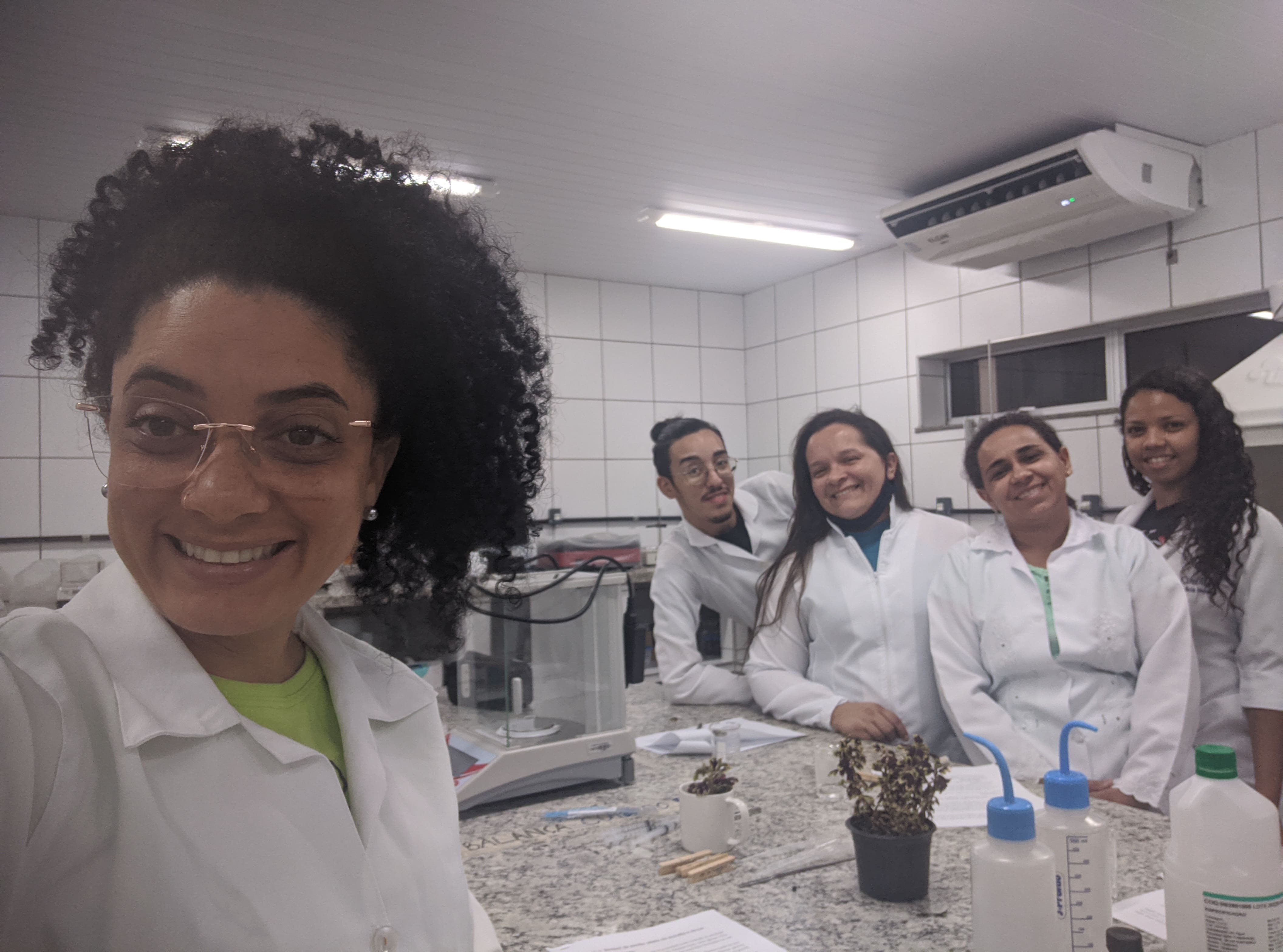 Uma mulher de óculos e cabelos cacheados está em primeiro plano, enquanto há quatro pessoas em segundo plano. Todas estão sorrindo, dentro de um laboratório