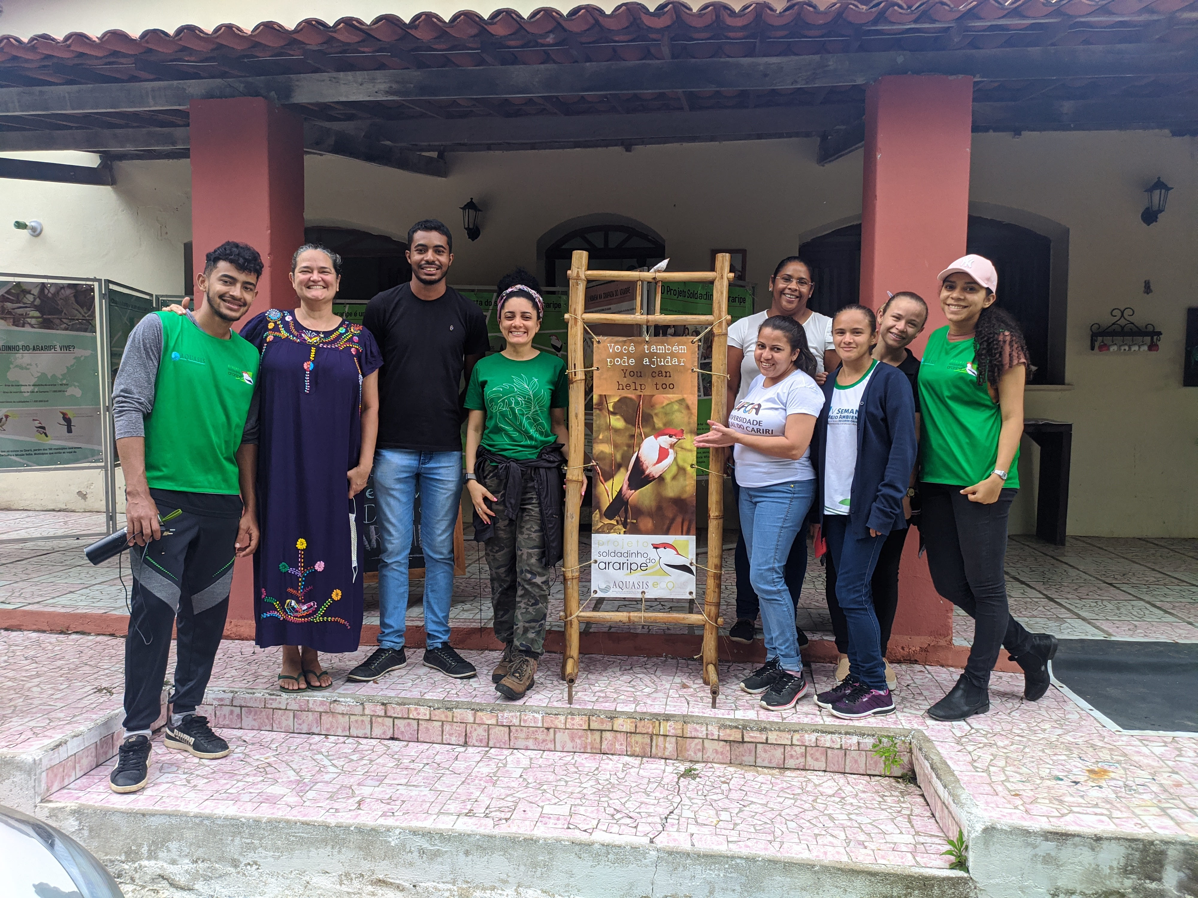 Foto de um grupo de pessoas (estudantes e professoras) sorrindo. No meio delas está um banner com uma imagem do soldadinho do Araripe