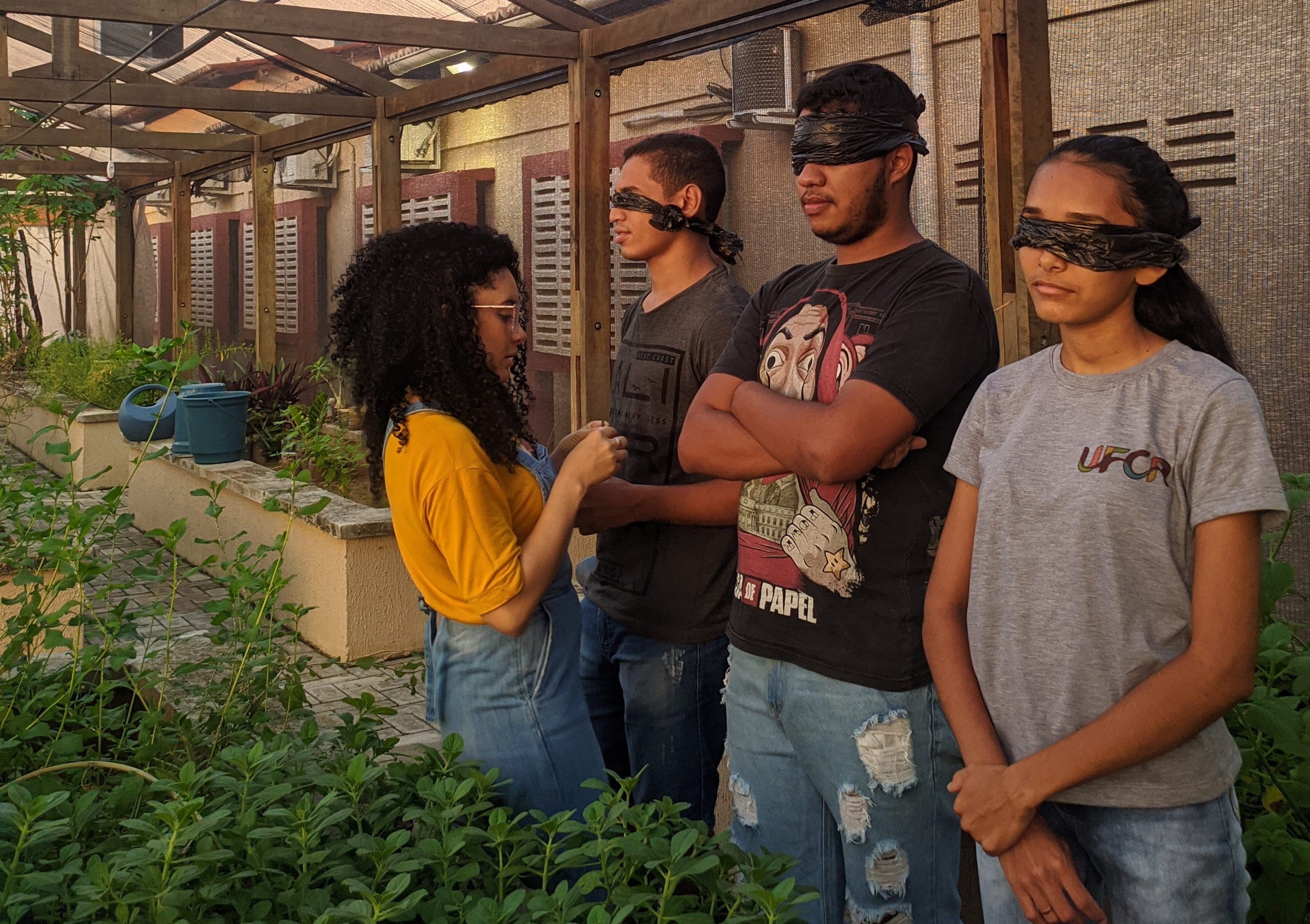 Três pessoas com os olhos cobertos por uma venda preta. Em frente a elas está uma outra pessoa segurando uma planta. Todos estão dentro de um espaço que cultiva várias plantas