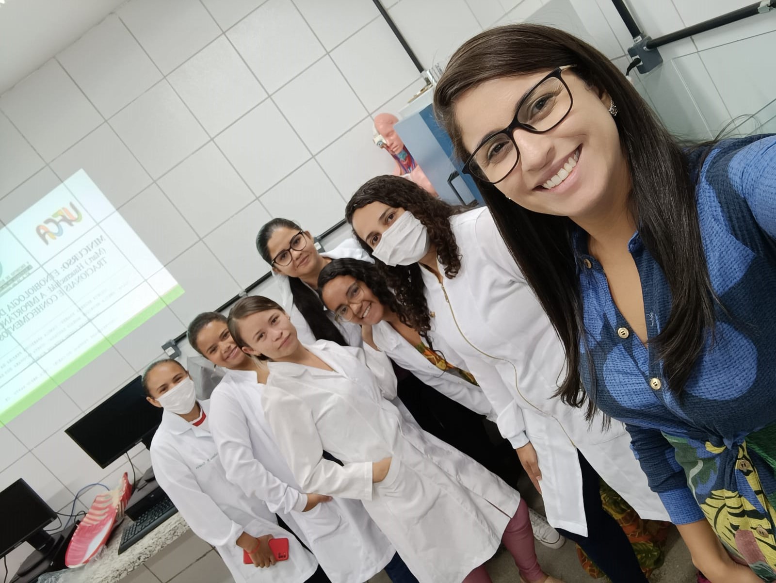 Uma mulher, de óculos, em primeiro plano fazendo uma selfie e outras seis mulheres, de jaleco, em segundo plano. Elas estão dentro de um laboratório