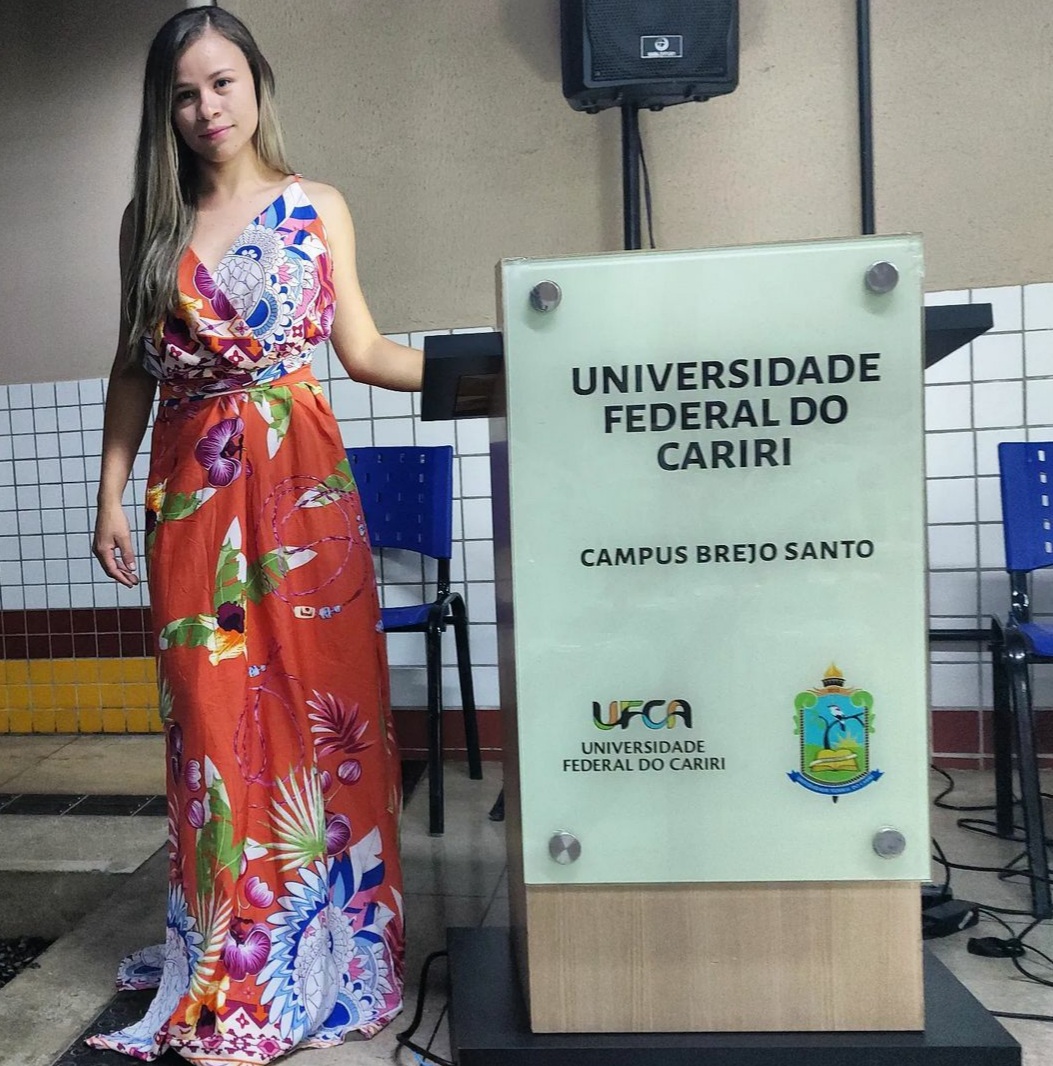 Um mulher de vestido colorido está em pé, ao lado de um púlpito, one está escrito Universidade Federal do Cariri - Campus Brejo Santo