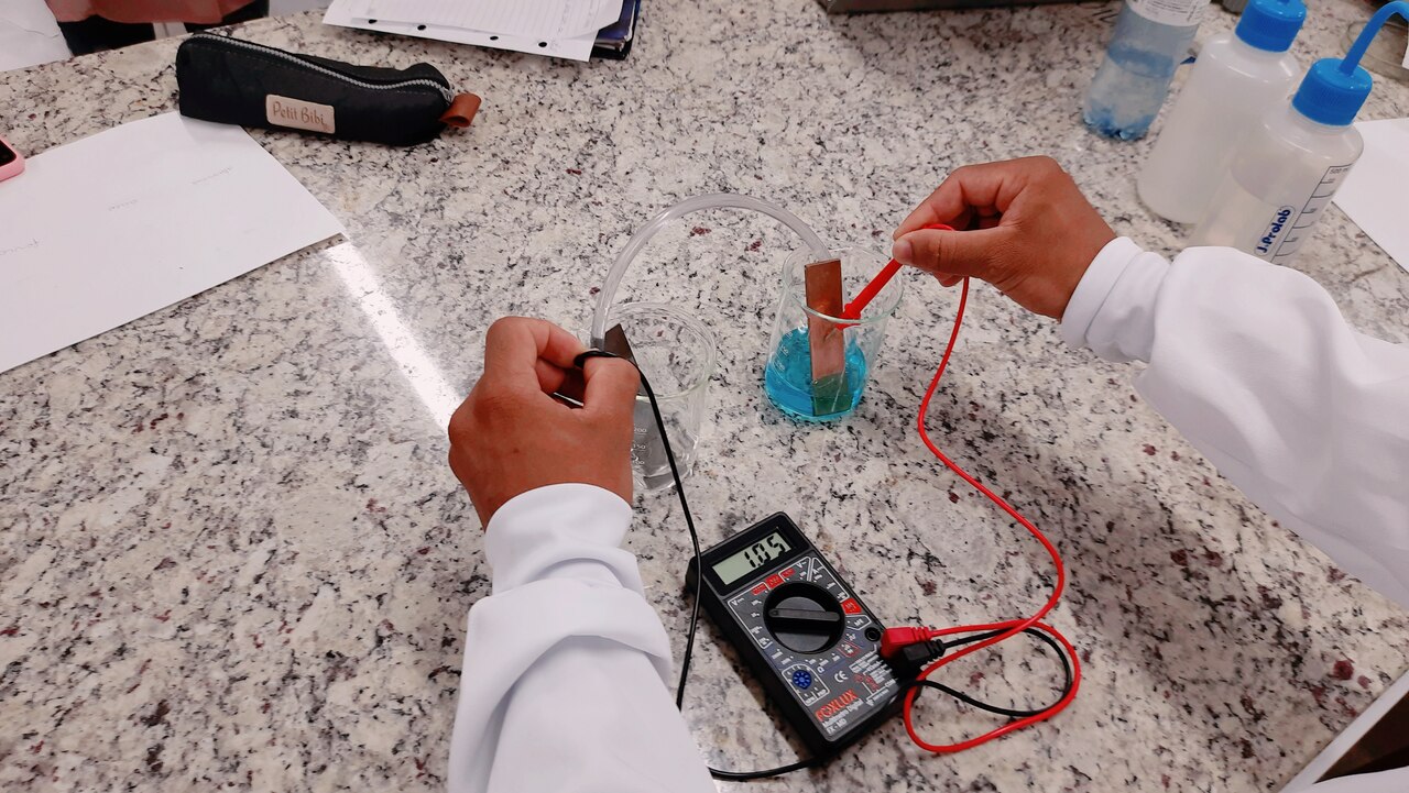 Mãos de um estudante segurando um equipamento, fazendo um experimento, na bancada do laboratório