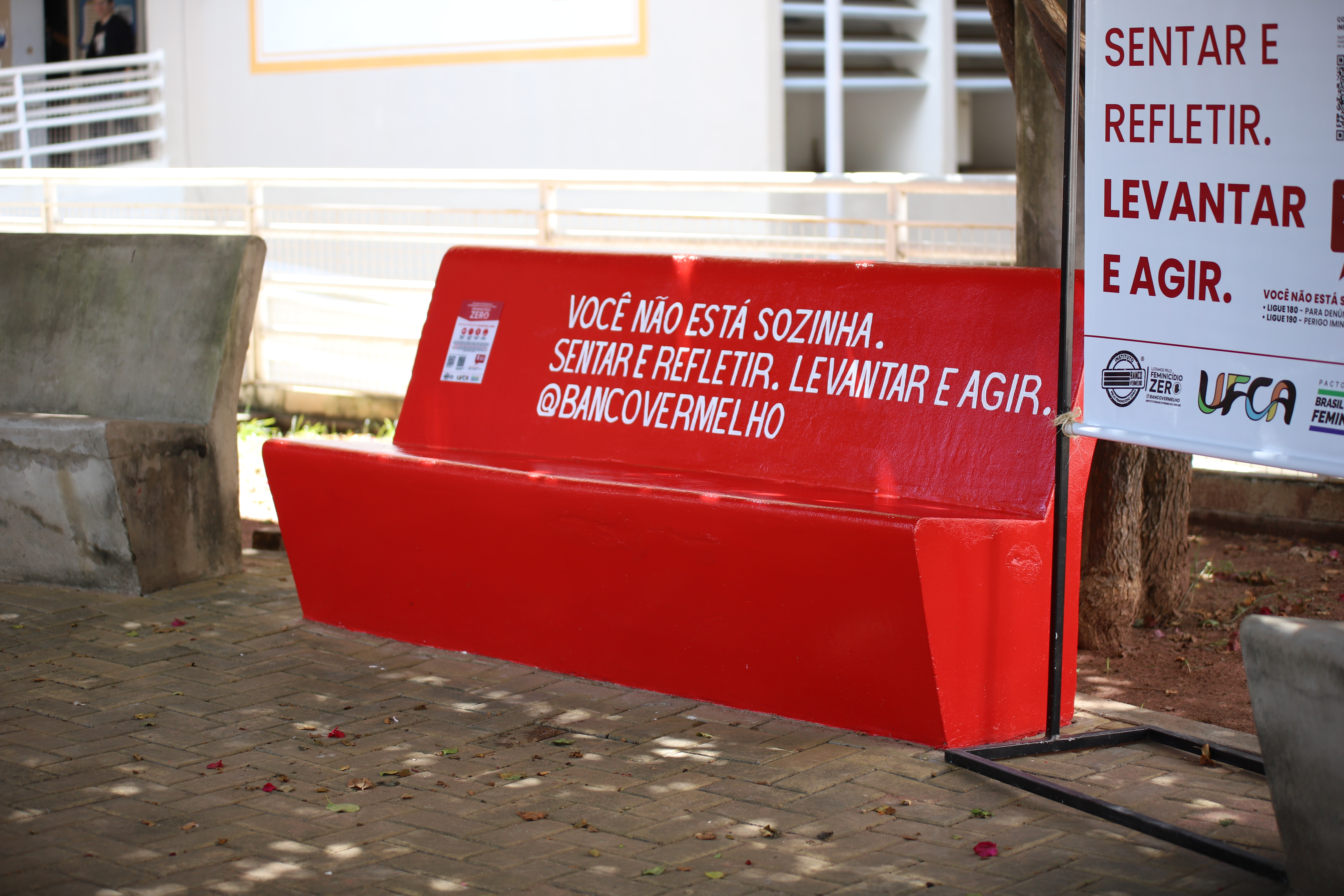 Banco de concreto pintado de vermelho em área externa do campus Juazeiro do Norte da UFCA. No encosto, há a frase “Você não está sozinha. Sentar e refletir. Levantar e agir. @bancovermelho”. Ao lado, aparece uma placa vertical da campanha Banco Vermelho.