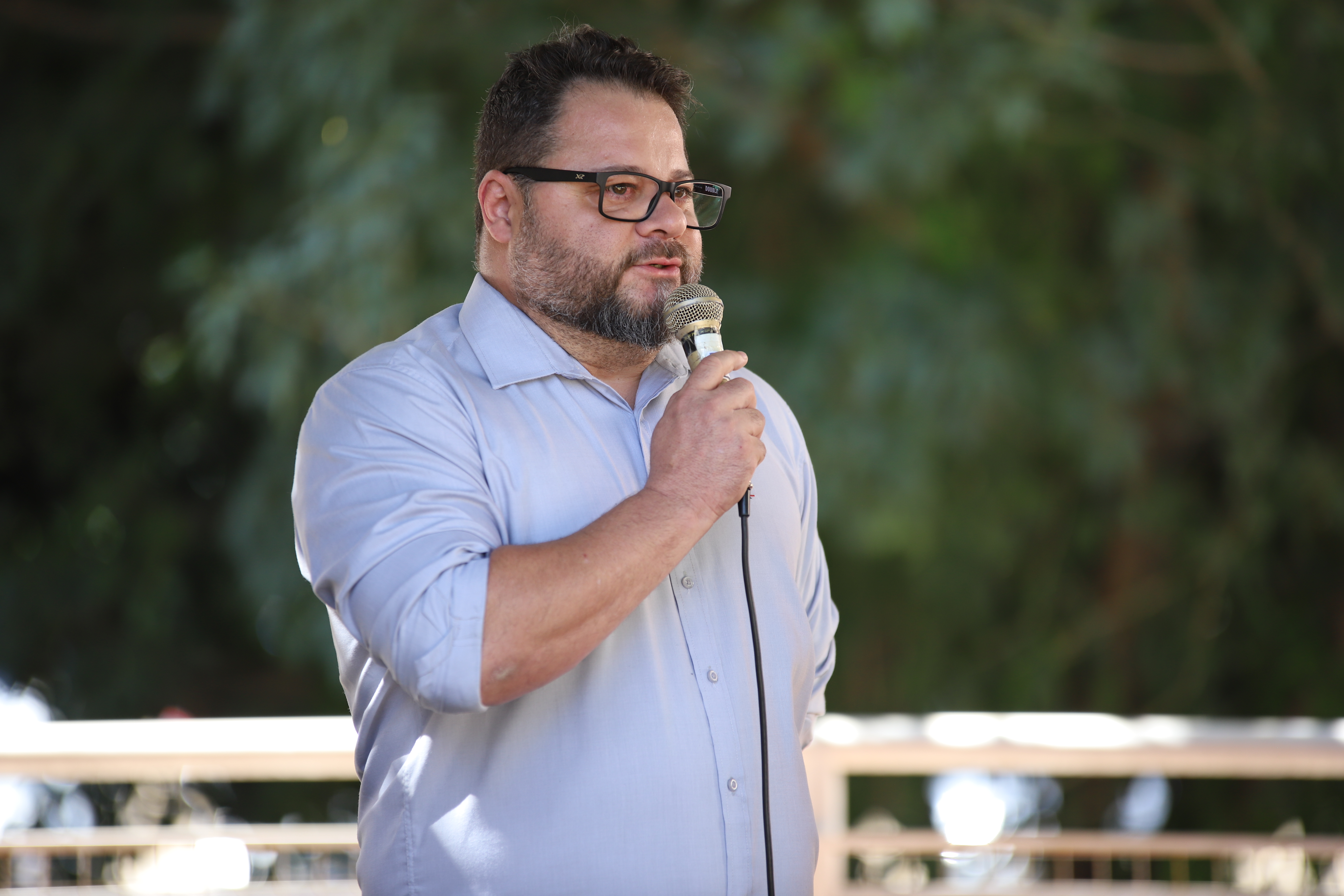 Homem de óculos, com camisa azul-clara, fala ao microfone em área externa da UFCA durante o ato de adesão à campanha Banco Vermelho. Ao fundo, a imagem mostra vegetação desfocada.