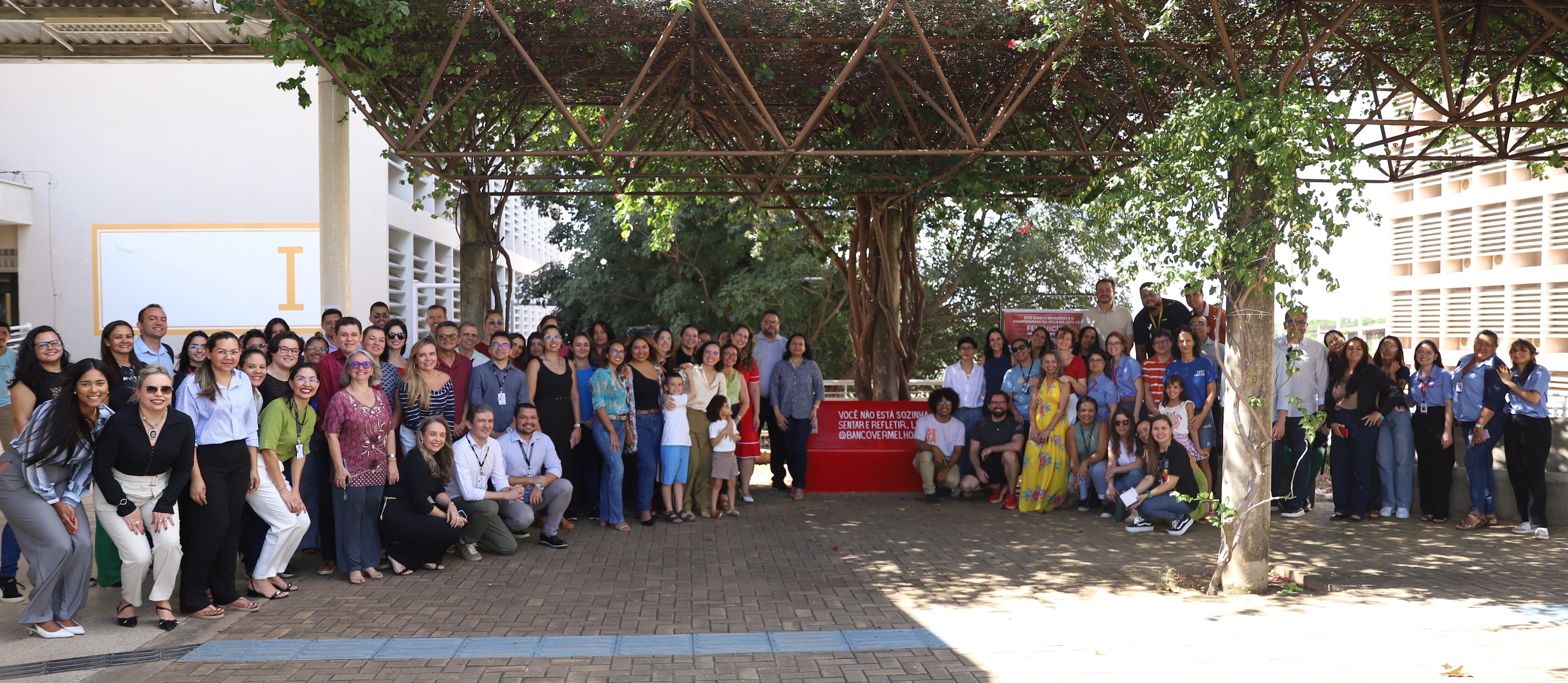 Grupo de pessoas reunido em área externa do campus Juazeiro do Norte da UFCA, durante o ato de adesão à campanha Banco Vermelho. Ao fundo, aparece o banco vermelho instalado como símbolo de enfrentamento à violência contra a mulher.