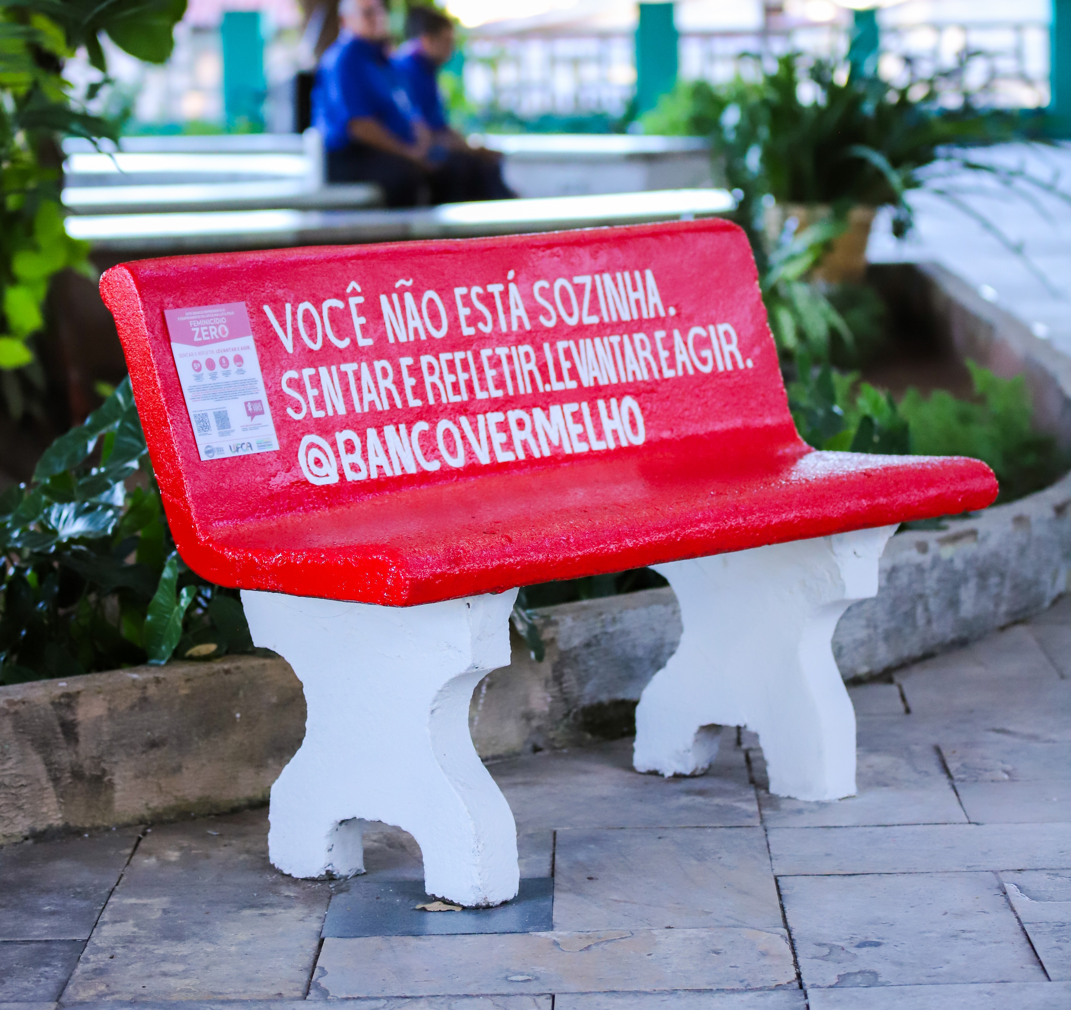 Banco vermelho instalado em área externa arborizada do campus Barbalha da UFCA. No encosto, há a frase: “Você não está sozinha. Sentar e refletir. Levantar e agir.”.