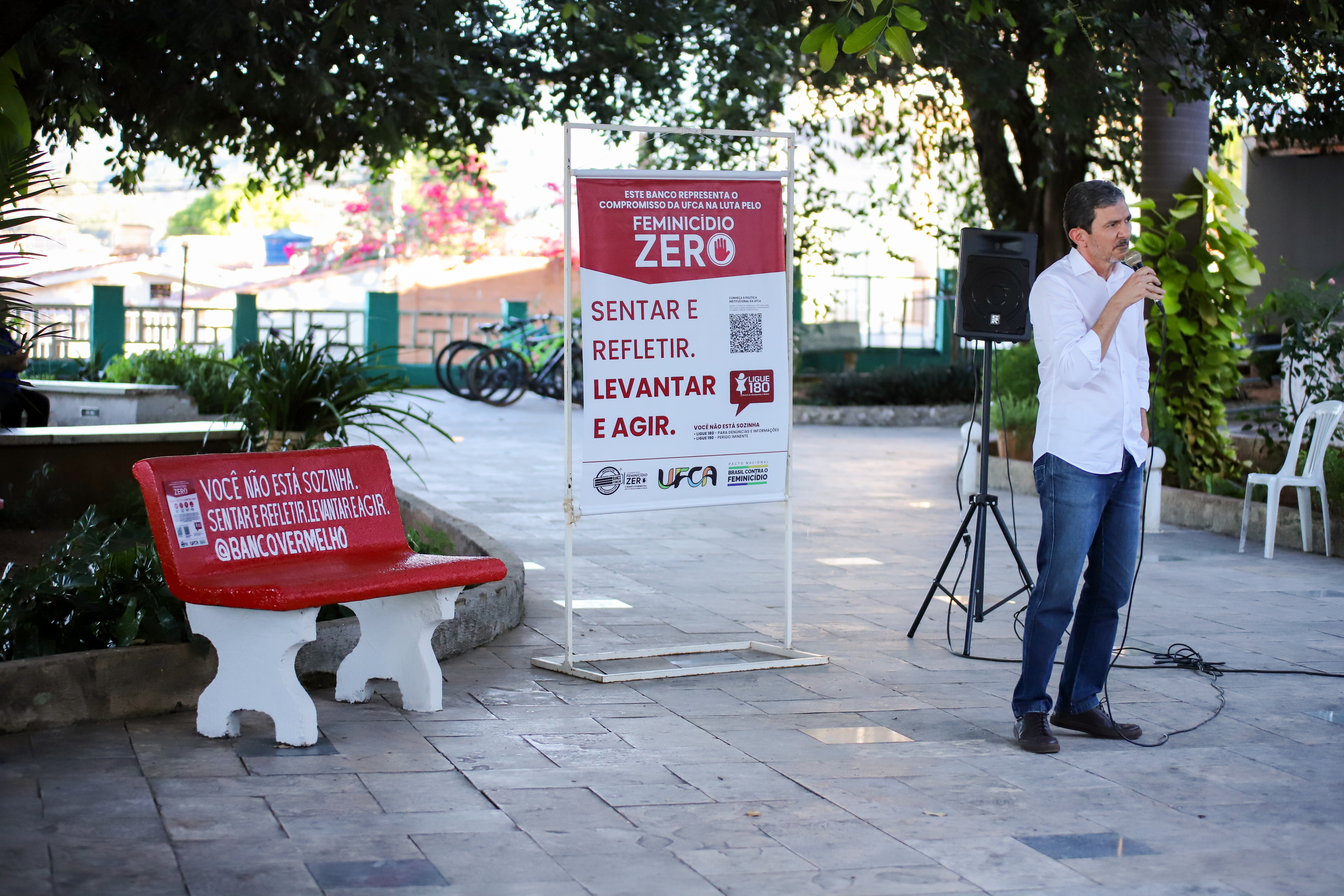 Professor Mário Pacheco, pró-reitor de Gestão de Pessoas da UFCA, fala ao microfone durante ação do Banco Vermelho no campus Barbalha. Ao lado, aparecem o banco vermelho e um banner da campanha com mensagens de enfrentamento à violência contra as mulheres.